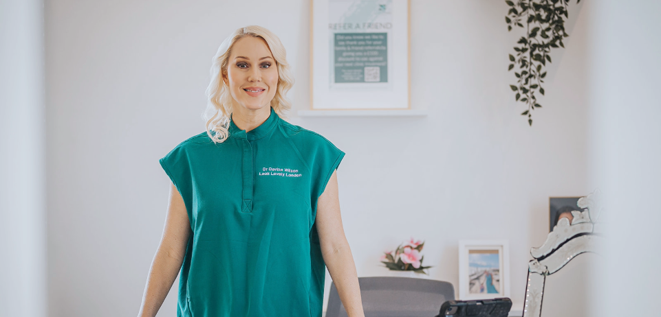 Dr Davina wearing a teal shirt smiles while standing in a waiting room with framed pictures and a plant on the wall.